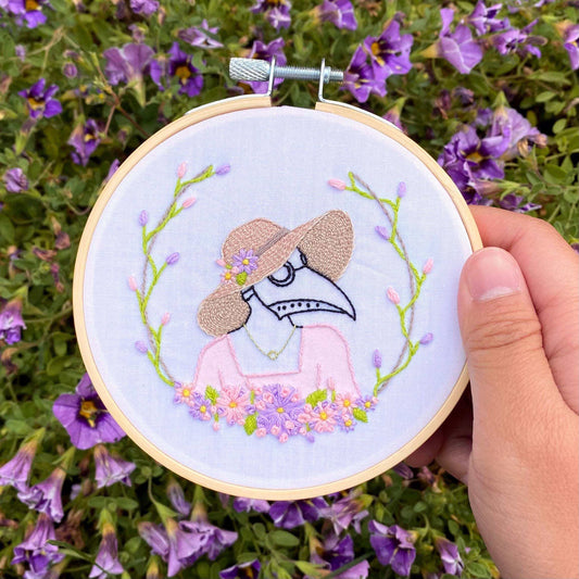 Hand embroidery kit with pastel plague doctor design, perfect for beginner/intermediate crafters; includes 4" hoop, fabric, thread, needles, paint, and tutorials.