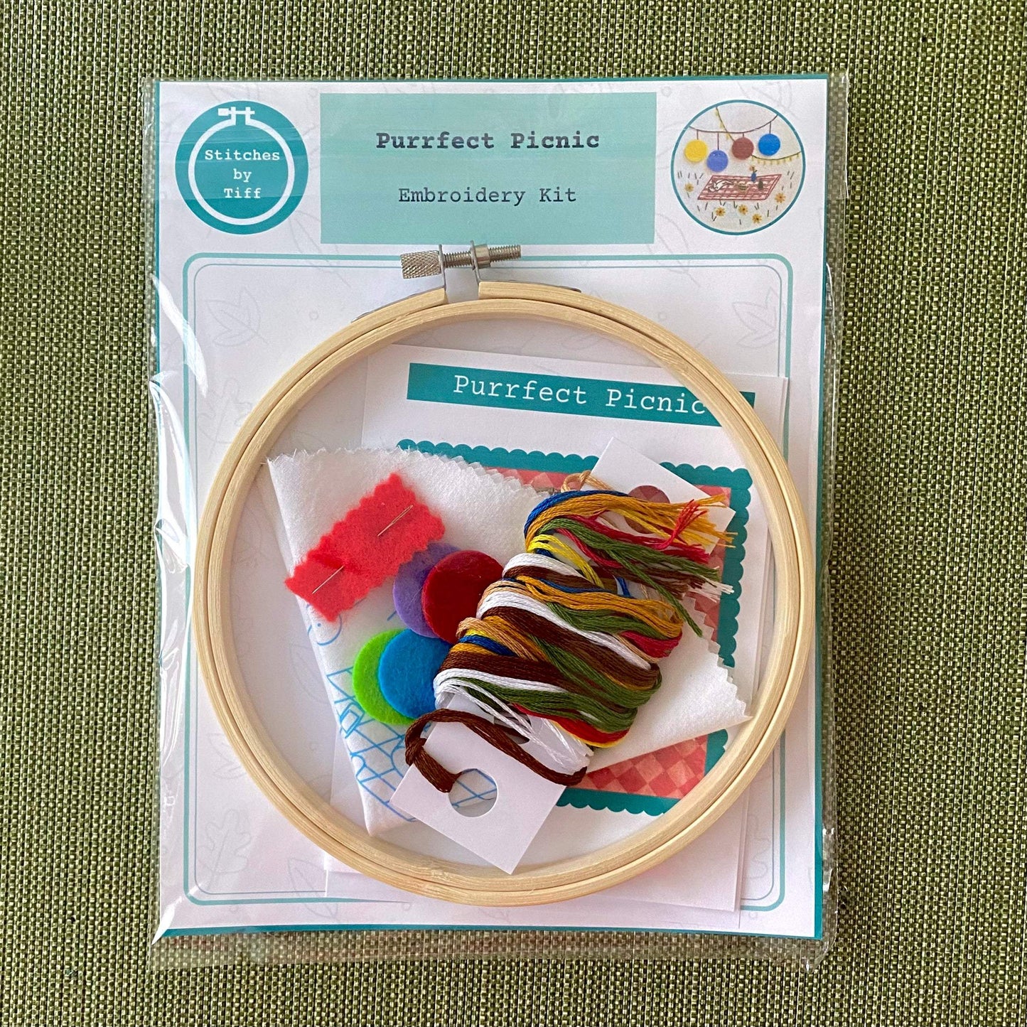 Purrfect Picnic Embroidery Kit with hoop, threads, and felt circles on a green background.
