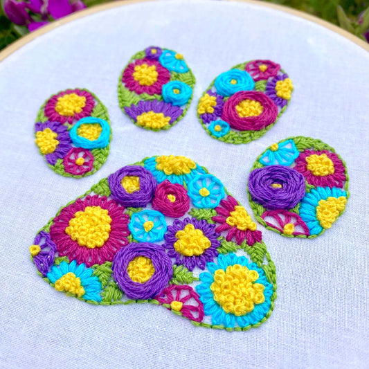 Colorful paw print embroidery design with flowers, showcasing various stitching techniques on white fabric in a bamboo hoop.